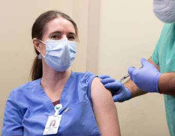 A woman wearing scrubs and a face mask receives a shot from a man in scrubs and a mask.