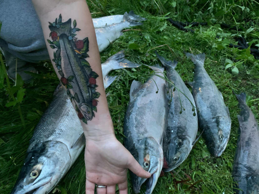 An arm with a salmon tattoo. The salmon is on a bed of roses. A pile of salmon in grass is in the background.