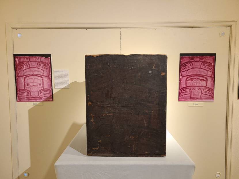 A museum display with an old, brown wooden box flanked by printed infrared images of formline paintings