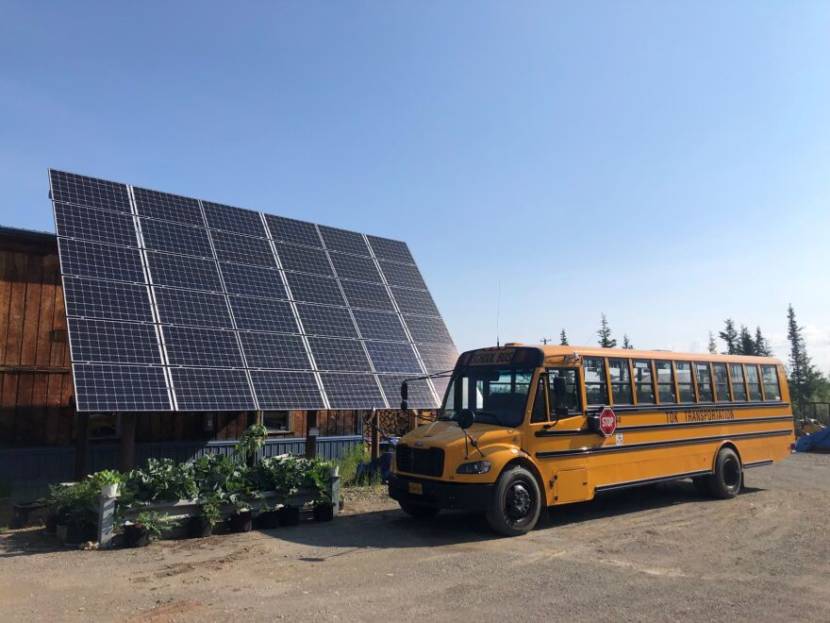 The electric school bus parked in front of a large bank of solar panels, about twice as tall as the bus