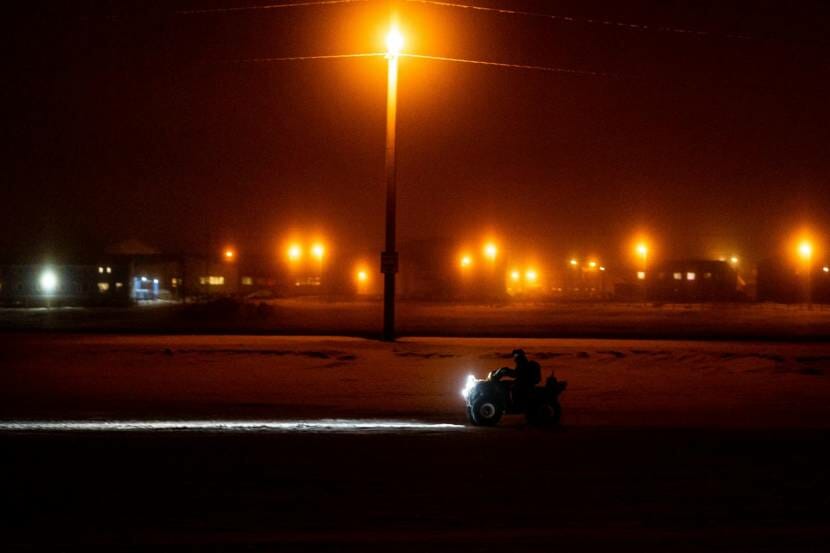 A 4-wheeler driving at night in the snow with homes and orange-glowing street lights in the background