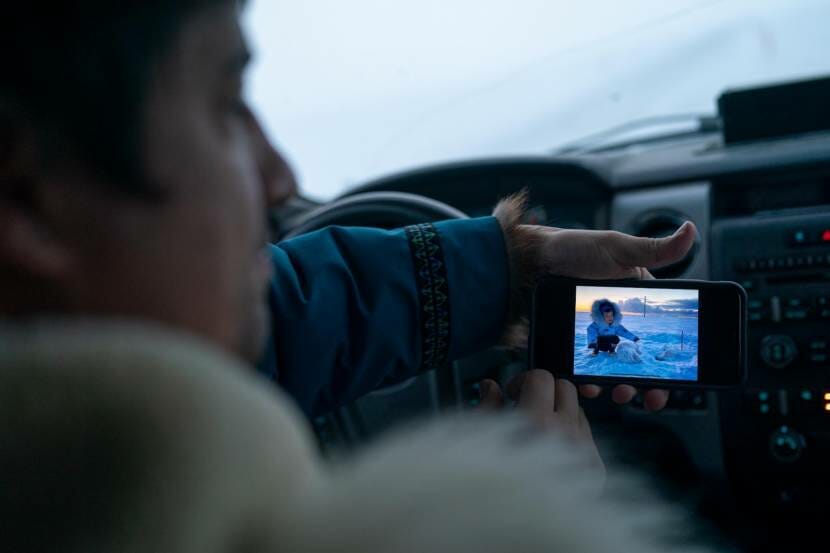 A man behind the wheel of a car holding out a cell phone that's displaying a picture of a child playing with a white fox in the snow
