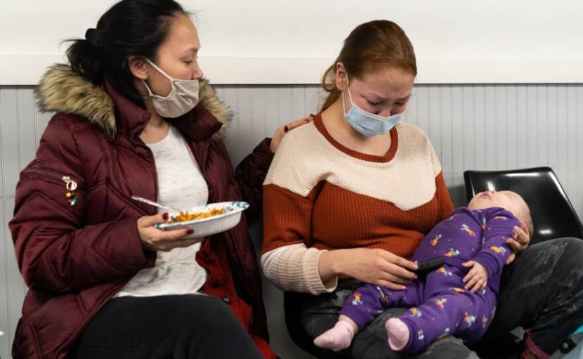 A woman puts her hand on the shoulder of another woman who is crying with a baby on her lap. 