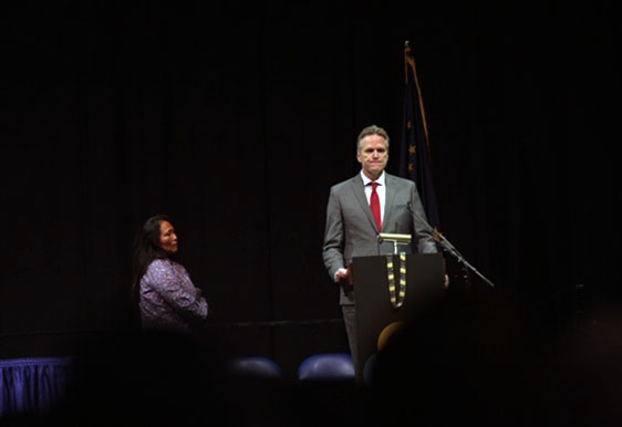 The governor, spotlit, standing behind a podium on a stage with a woman to his right