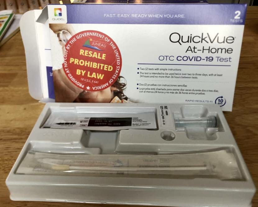 An opened box containing an at-home COVID-19 test. A plastic tray inside includes a long nasal swab, a small tube with liquid, and a test strip in a plastic sleeve.