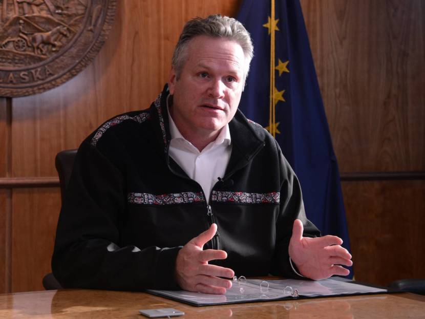 Gov. Dunleavy sits behind a desk wearing a Copper River fleece jacket, with the Alaska state flag behind him