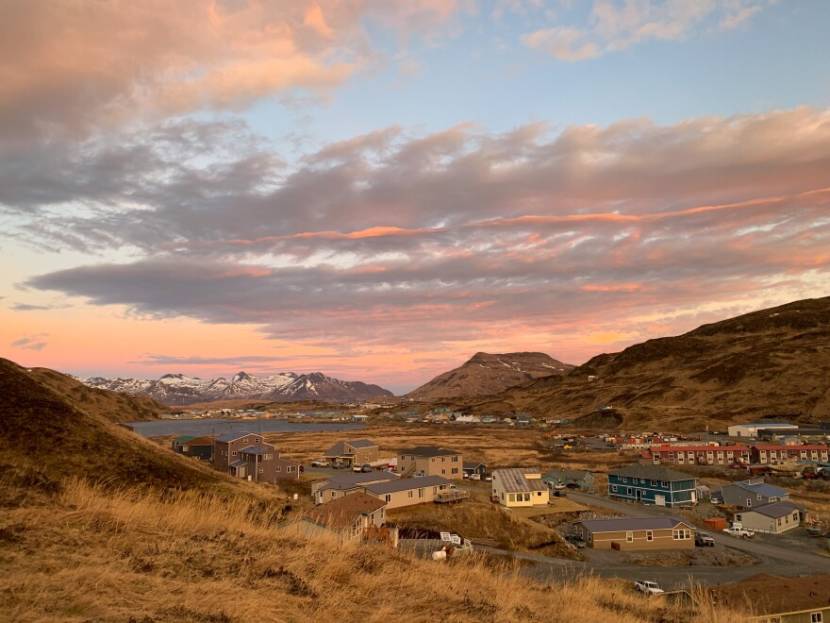 Scattered buildings in a setting of brown hills and faraway snowcapped mountains at sunset.