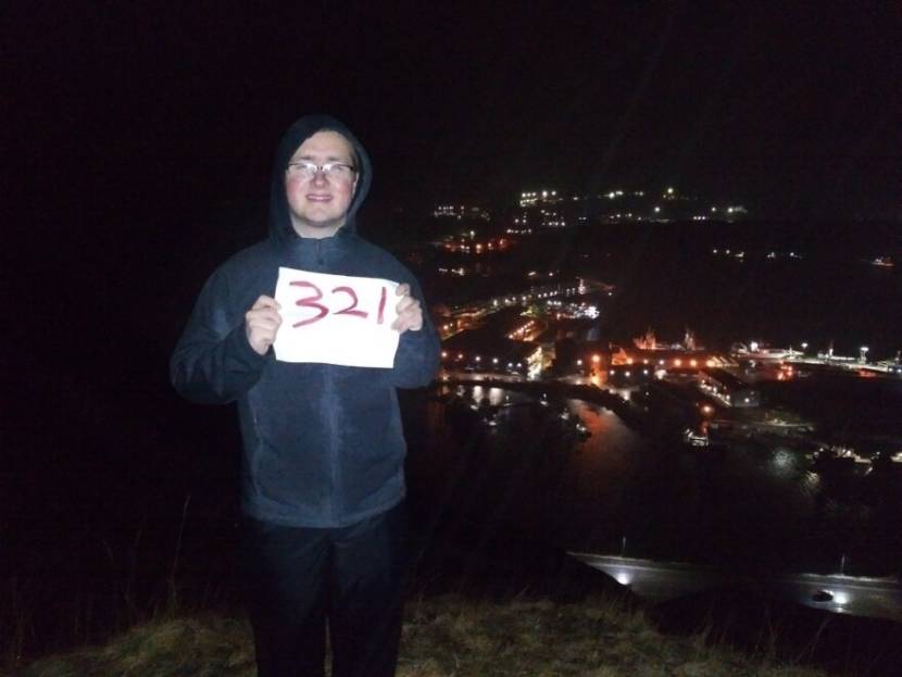 A man standing on top of a hill at night holding a sign that says 321 on it. The lights of Dutch Harbor and Unalaska are in the background below.