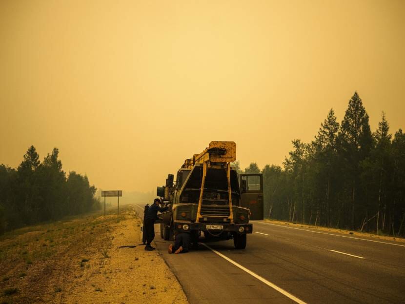 Two men working on a truck by the side of a road through a forest. The sky is yellow with wildfire smoke.