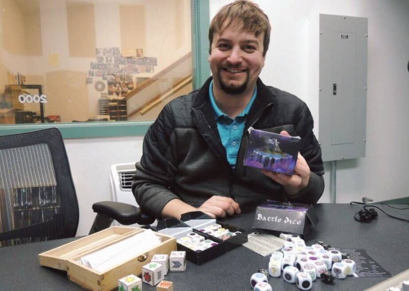 A man sitting at a table full of dice, holding up the box for Battle Dice