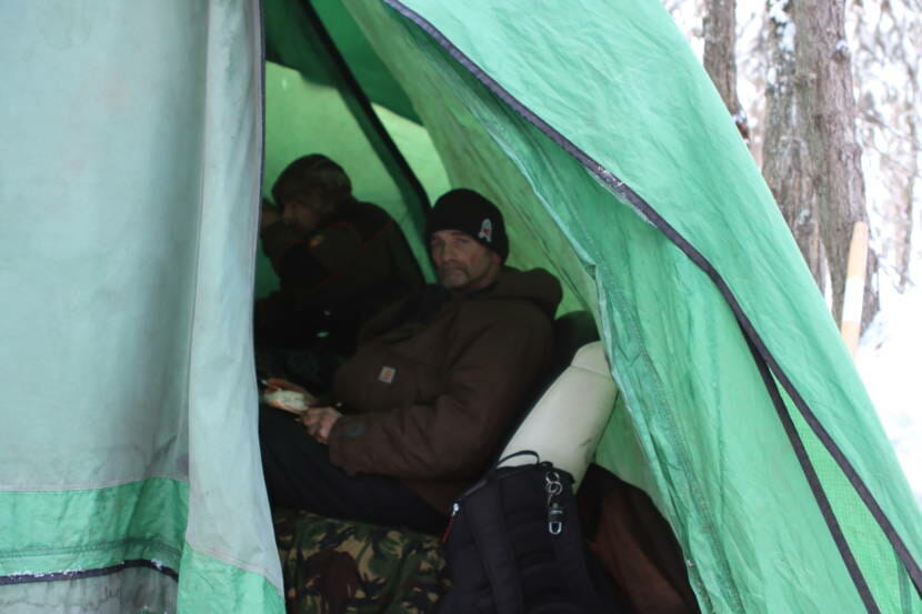 Two men sitting inside a green tent, photographed from outside through the tent's entrance.