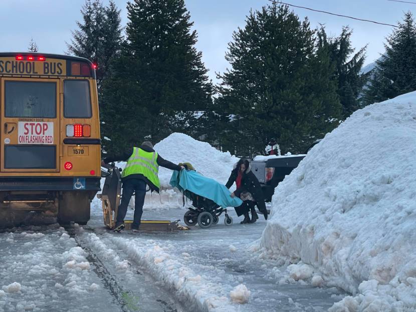 People moving a woman in a wheelchair onto a school bus's wheelchair lift, in wintry conditions