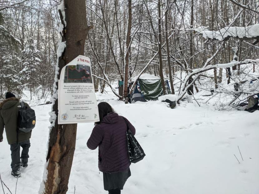 Two people walk through the snow towards a camp, passing a notice on a tree saying the camp will be removed.