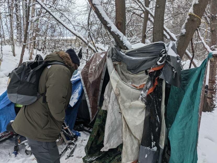 A man in winter clothes approaching a makeshift camp in the woods.