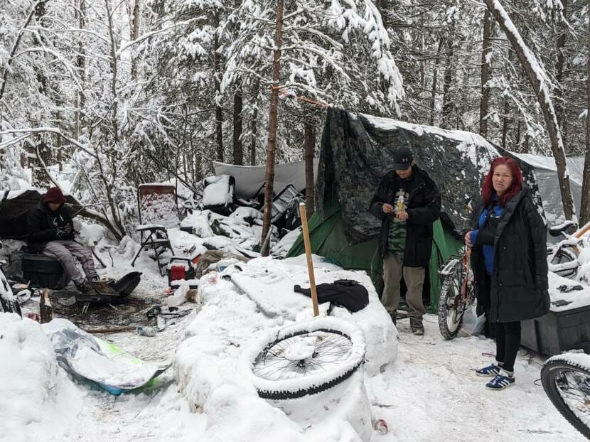 Three people at a small camp in the woods in the snow, with bicycles, chairs and other possessions outside.