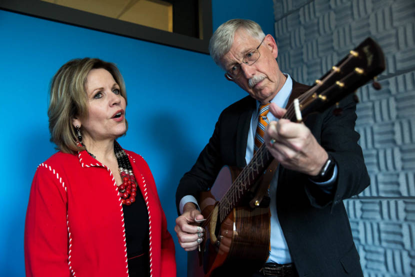 A man in a suit fingerpicking an acoustic guitar while a woman stands next to him, singing
