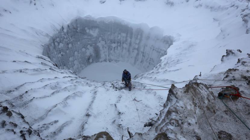 A man is roped up at the edge of a huge hole in the snowy tundra