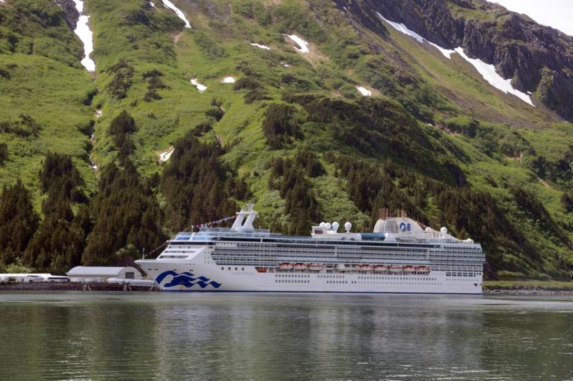 A cruise ship sailing in front of steep, green forested slopes.