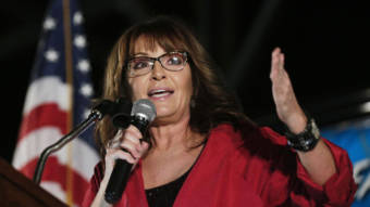 Sarah Palin holding a microphone and speaking in front of an American flag