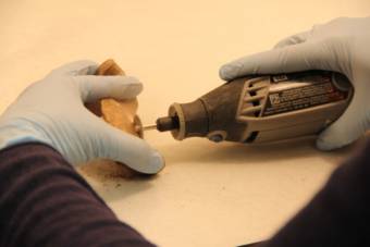 A pair of gloved hands beginning to saw into a bone fragment with a dremel tool