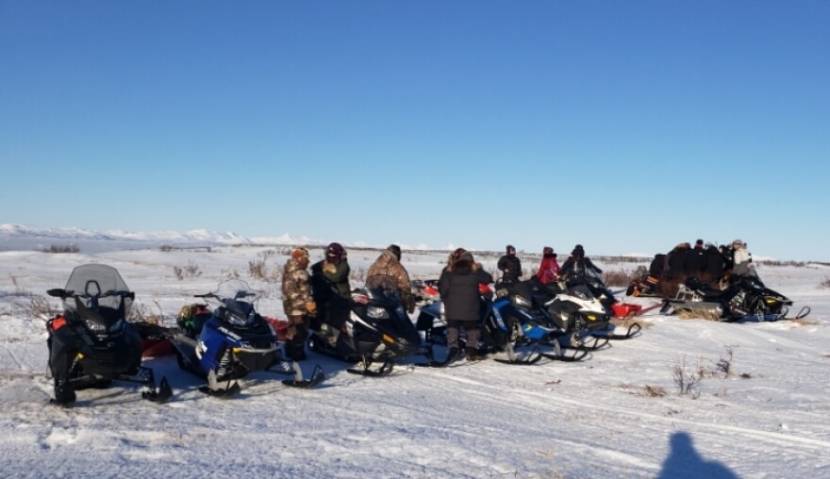 People standing around a line of about 10 snowmachines on the open tundra