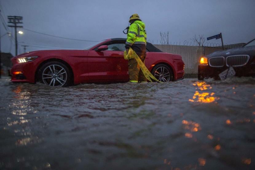 A firefighter holding caution tape stands at the driver's side window of a red sports car that's up to its rims in water on a flooded street