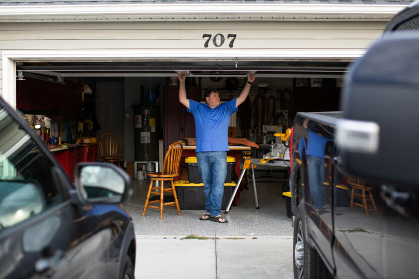 A man holds a garage door open to show a home workshop inside.