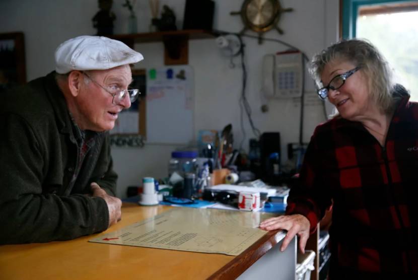 An older man in a flat cap speaks to a woman from across a counter