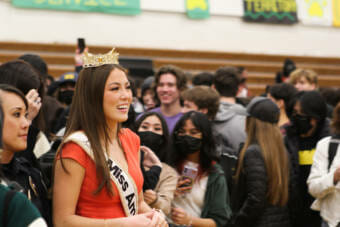 A woman wearing a crown and Miss America sash stands in a crowded gymnasium