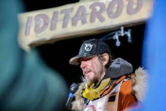 A musher with an icy beard stands under a sign that says "Iditarod"