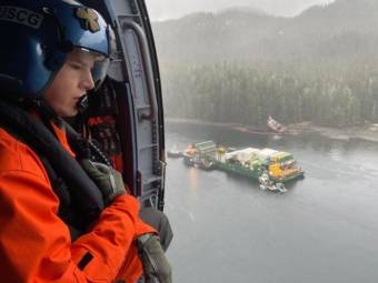 A Coast Guard member looks out the door of a helicopter at a container barge surrounded by other vessels off a wooded coast