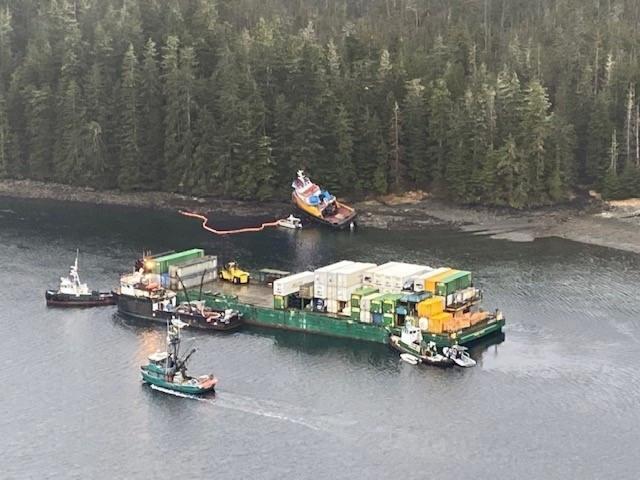 A container barge is surrounded by smaller vessels while a tugboat lays pushed up on the shore