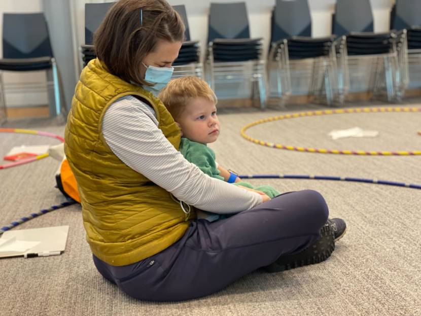 A woman in a mask sits on the floor with a small child in her lap at a library during storytime