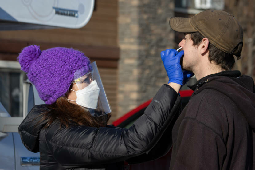 A woman wearing a purple toque and a face shield sticks a nasal swab up a man's nose, outdoors