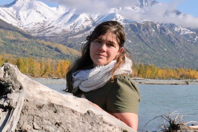 A portrait of a woman standing outside by a river, with snowy mountains behind her