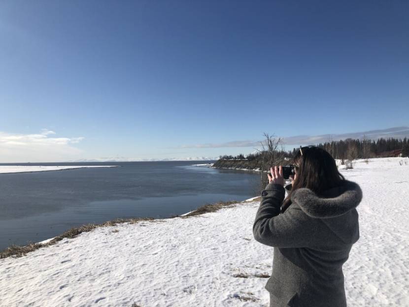 A woman on a snowy shore looks out on the water with binoculars