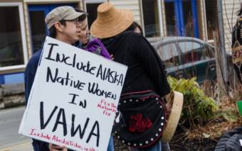A man in a small group of people holds a sign that says "Include Alaska Native women in VAWA)