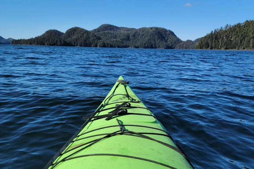 The front of a neon green sea kayak on the water, with forested shores in the distance