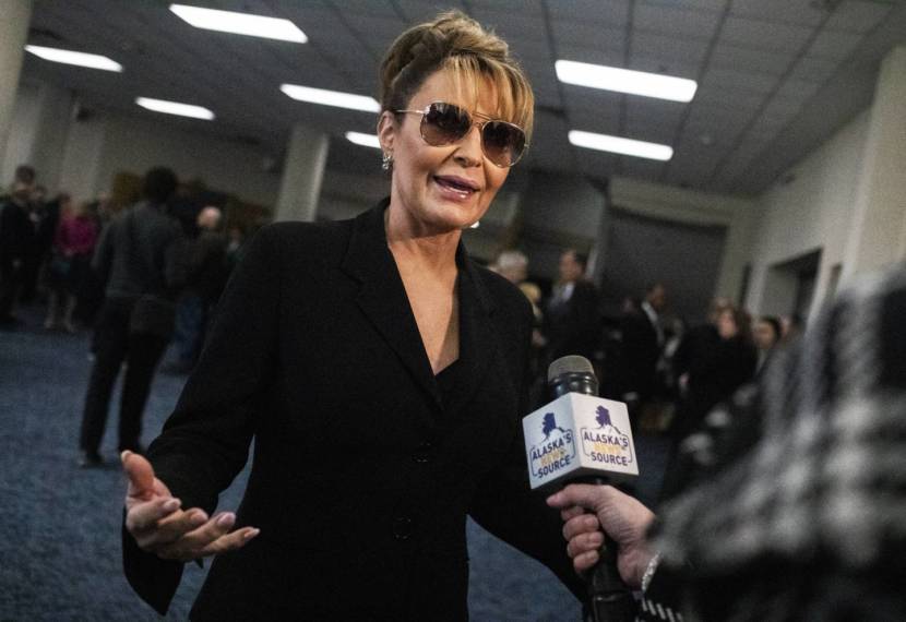 Sarah Palin, wearing sunglasses indoors, speaks into a microphone