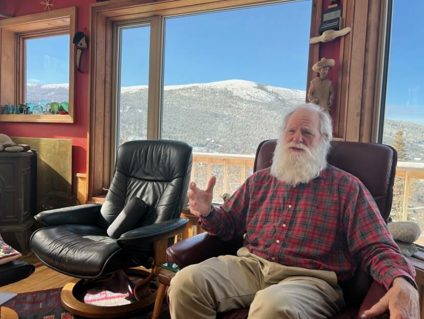 A man with a large, white beard sitting in a home with sweeping mountain views