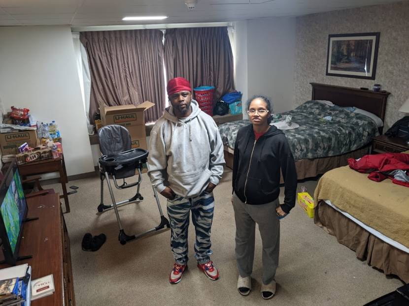 A couple stands in the middle of a hotel room filled with their belongings.