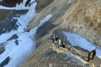 A group of workers stand at the edge of a steep drop in a gravel road along a mountainside