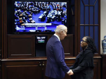 President Biden clasping hands with Ketanji Brown Jackson. A large screen behind them shows the Senate vote.