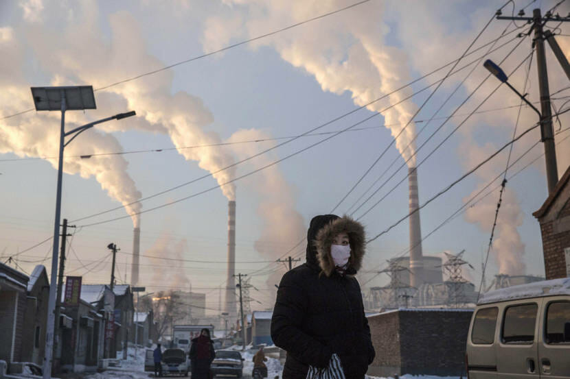 A masked person in a parka walking in a city with many tall stacks emitting white smoke in the background