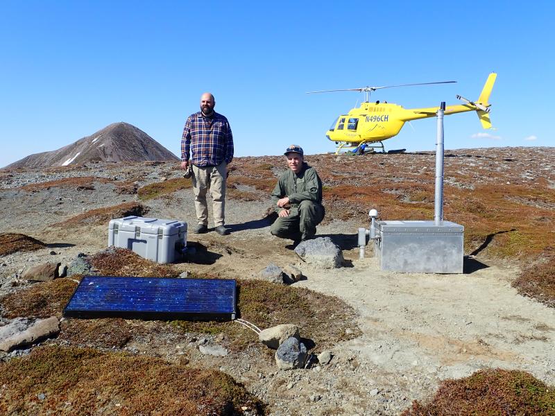 Two men pose with scientific equipment on a treeless ridge with a yellow helicopter behind them