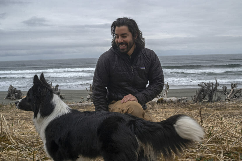 A man squats next to a border collie-looking dog on a beach