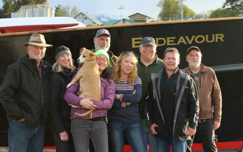 A group photo of eight people and a dog on a dock in front of the Endeavour
