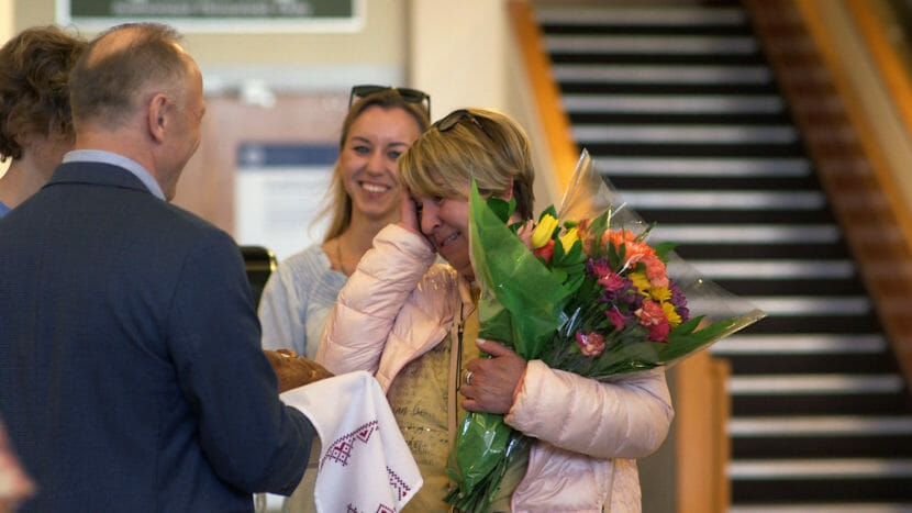 A woman in an airport holds one hand to her face while holding a bouquet of flowers