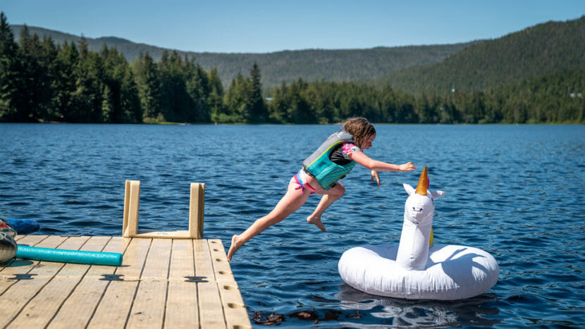 A child in a life jacket jumps off a dock onto a unicorn pool float in a lake