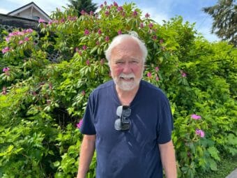 An older man standing in front of a shrub with two pairs of glasses hanging from his t-shirt collar